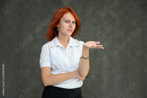 Photo Portrait of a cute girl woman with bright red hair manager in a white shirt on a gray background in the studio. He talks, shows his hands in front of the camera with emotions.