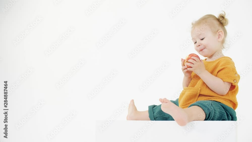 Baby wearing orange and green clothes eats apple against white background Baby wearing orange and green clothes eats apple against white background
