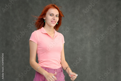 Photo Portrait of a cute woman girl with bright red hair in a peach t-shirt and pink skirt on a gray background in the studio. He talks, shows his hands in front of the camera with emotions.