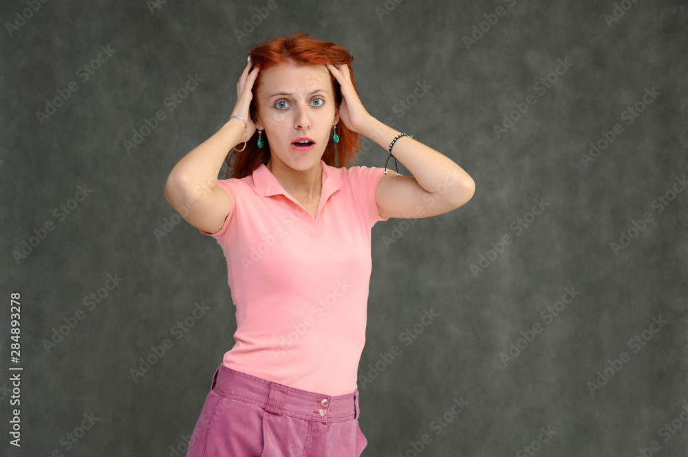 Photo Portrait of a cute woman girl with bright red hair in a peach t-shirt and pink skirt on a gray background in the studio. He talks, shows his hands in front of the camera with emotions.