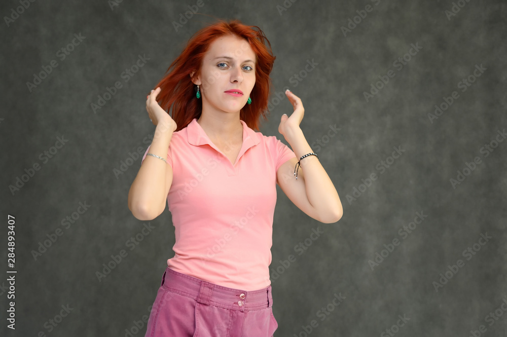 Fototapeta premium Photo Portrait of a cute woman girl with bright red hair in a peach t-shirt and pink skirt on a gray background in the studio. He talks, shows his hands in front of the camera with emotions.