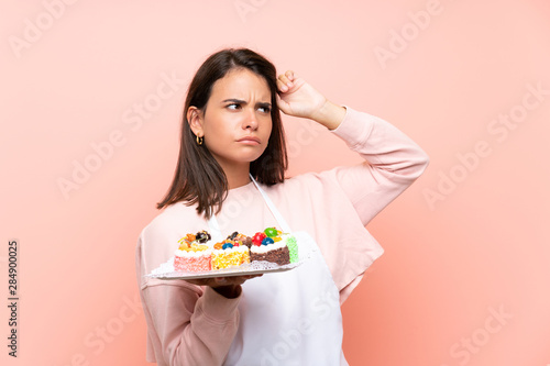 Young girl holding lots of different mini cakes over isolated background having doubts and with confuse face expression