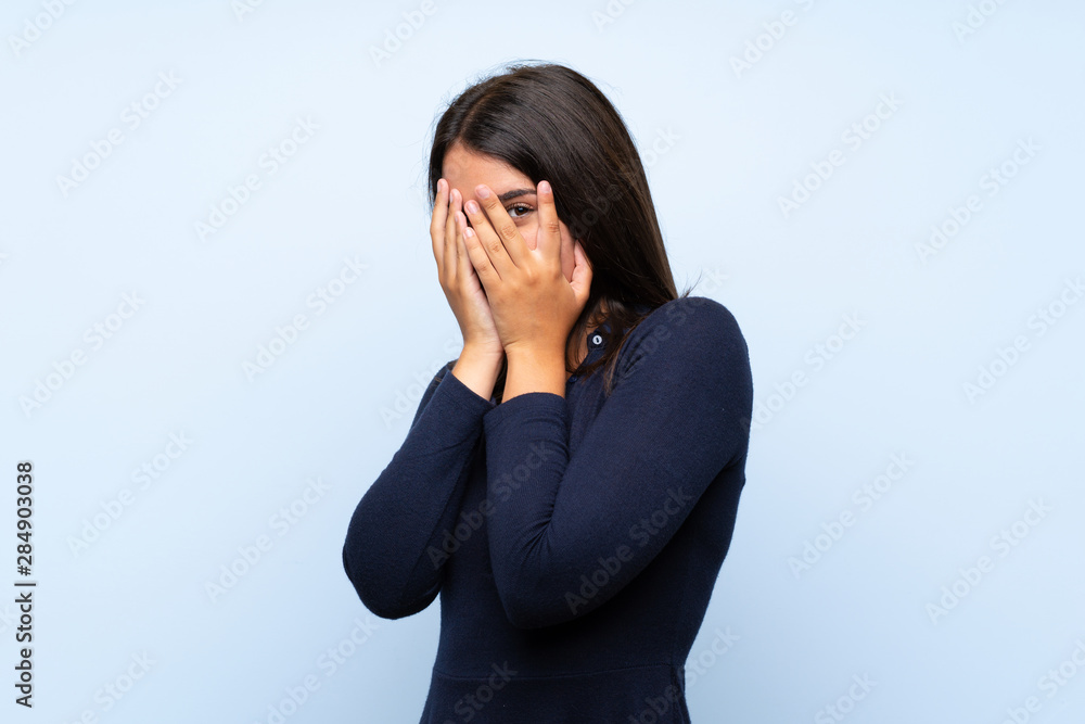 Young girl over isolated blue background covering eyes and looking through fingers Young girl over isolated blue background covering eyes and looking through fingers
