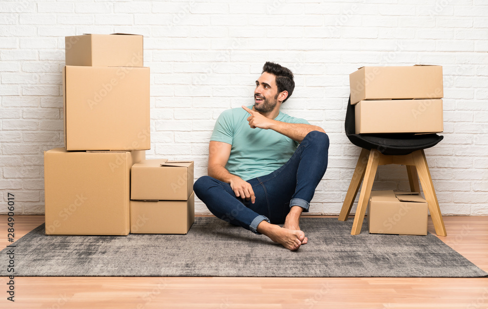 Handsome young man moving in new home among boxes pointing finger to the side Handsome young man moving in new home among boxes pointing finger to the side