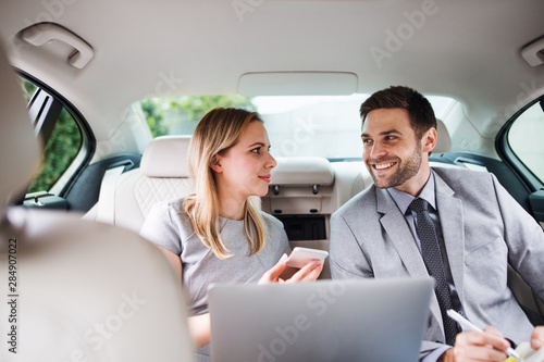Business couple with laptop sitting on back seats in car, talking.