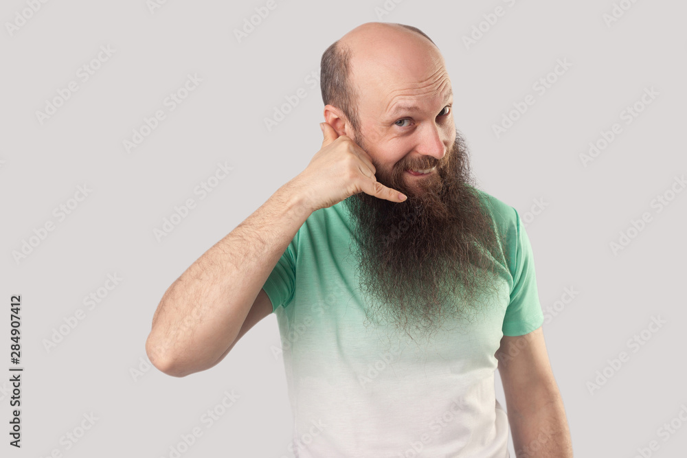 Call me! Portrait of happy middle aged bald man with long beard in light green t-shirt standing, looking with call hand gesture and asking to call him. indoor studio shot, isolated on grey background Call me! Portrait of happy middle aged bald man with long beard in light green t-shirt standing, looking with call hand gesture and asking to call him. indoor studio shot, isolated on grey background