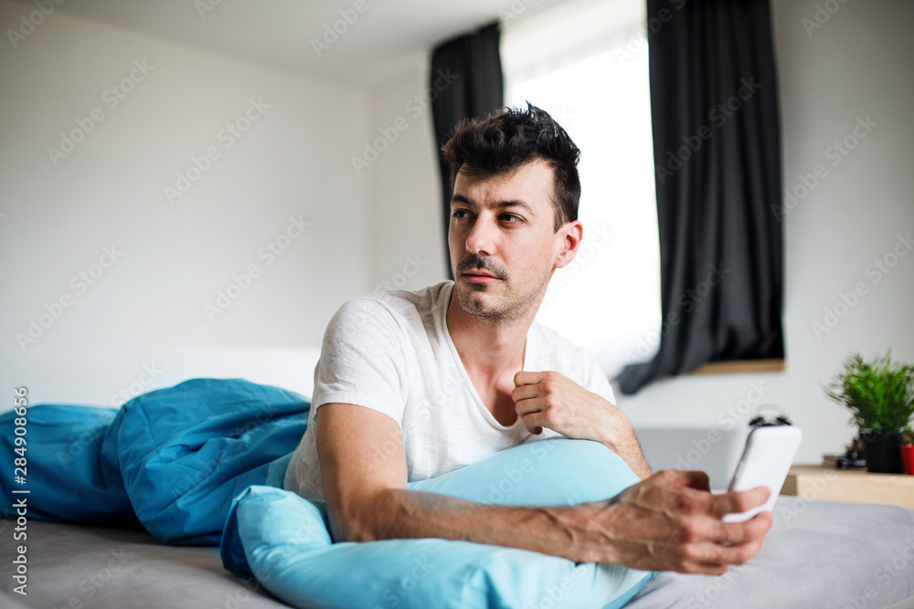 Young man with smartphone in bed at home, text messaging.
