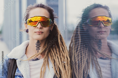 Beautiful styilish girl in big toned glasses, tattoo on the neck and piercing is posing near mirroed wall. photo