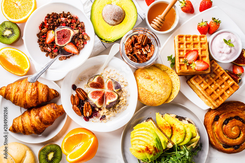 Breakfast table with avocado toast, oatmeal, waffles, croissants on white background.