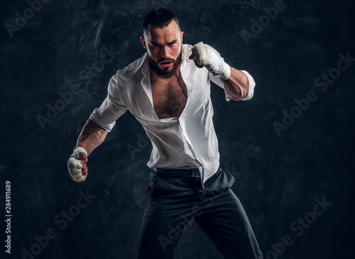 Agressive handsome man in white shirt is demonstraiting his punch at dark photo studio.