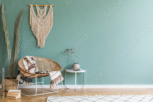 Stylish minimalistic interior of living room with design rattan armchair, palm leaf in basket, plaid, beige macrame on the wall, flowers and elegant accessories. Eucalyptus color of wall. Copy space. 