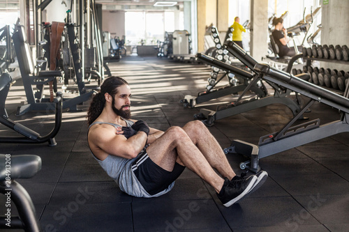 Portrait of young adult muscular built athlete with long curly hair working out in gym, lying on floor and swinging press with crossed arms on chest and bent legs. indoor, looking away