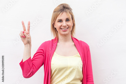 Portrait of happy beautiful blond young woman in yellow shirt and red blouse standing with victory or peace sign and looking at camera with toothy smile. studio shot, isolated on white wall background