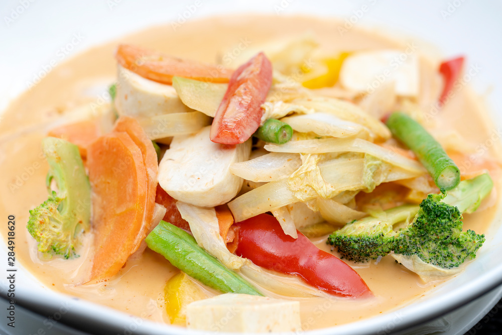 Mixed vegetables in creamy red curry coconut sauce with tofu in a white plate on a wooden table , traditional Thai food.