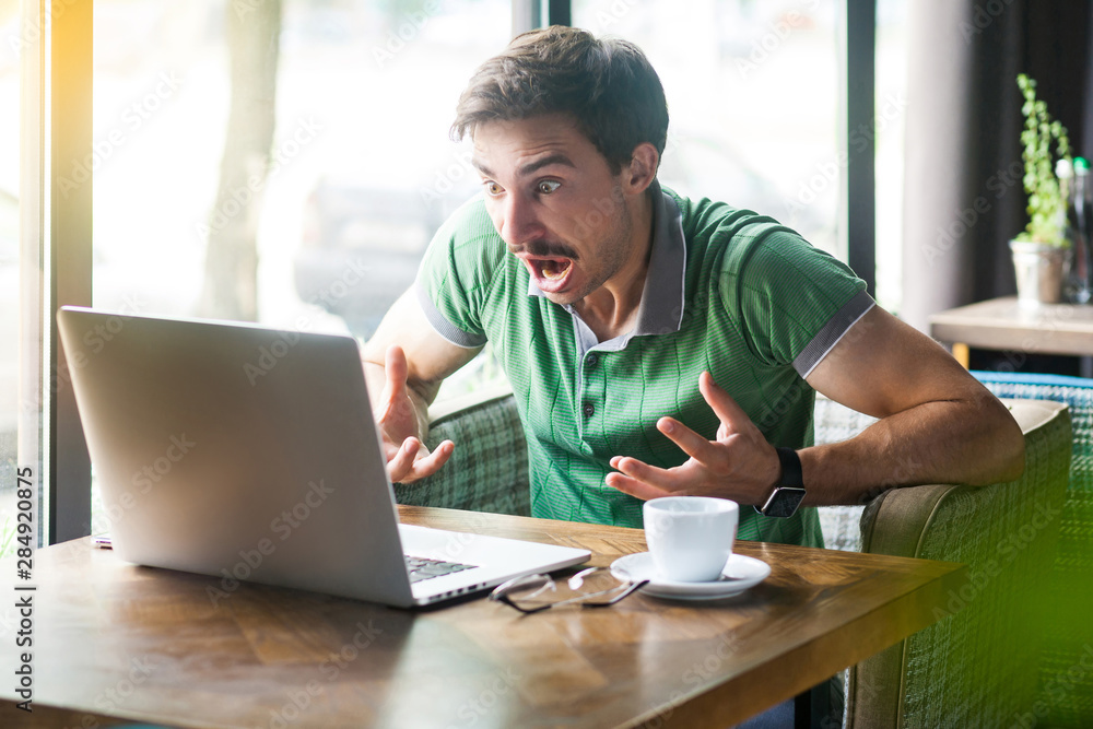 Young shocked businessman in green t-shirt sitting and looking at laptop display and reading bad news with unbelievable face expression. business concept. indoor shot near big window at daytime. Young shocked businessman in green t-shirt sitting and looking at laptop display and reading bad news with unbelievable face expression. business concept. indoor shot near big window at daytime.