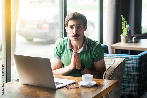 Please forgive me or help! Young worry businessman in green t-shirt sitting and looking at camera and pleading to help or forgive. business problem concept. indoor shot near big window at daytime.