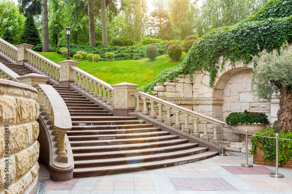 granite staircase with stone railings and balustrades with a wall rustic design in the background landscape with plants from bushes and trees and sunny glare. granite staircase with stone railings and balustrades with a wall rustic design in the background landscape with plants from bushes and trees and sunny glare.