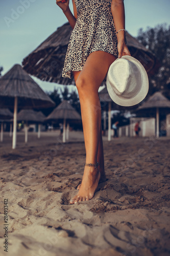 Young pretty girl in a dress walking on sandy beach in the evening while holding hat in her hand