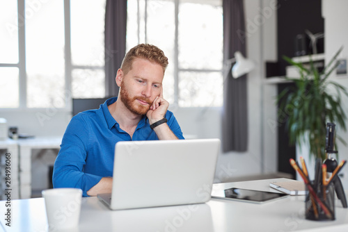 Young handsome Caucasian bearded employee leaning on desk and using laptop while sitting in modern office. Start up business concept.