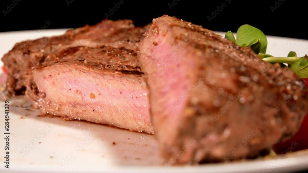A piece of fried meat garnished with Cherry Tomatoes and microgreen rotating on a white plate. Tasty grilled steak cut into two parts close-up. Demonstration of the finished dish. A piece of fried meat garnished with Cherry Tomatoes and microgreen rotating on a white plate. Tasty grilled steak cut into two parts close-up. Demonstration of the finished dish.