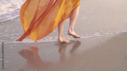 Female bare feet and fluttering dress at sunset. Young barefoot girlrunning in long orage dress along the surf line. photo