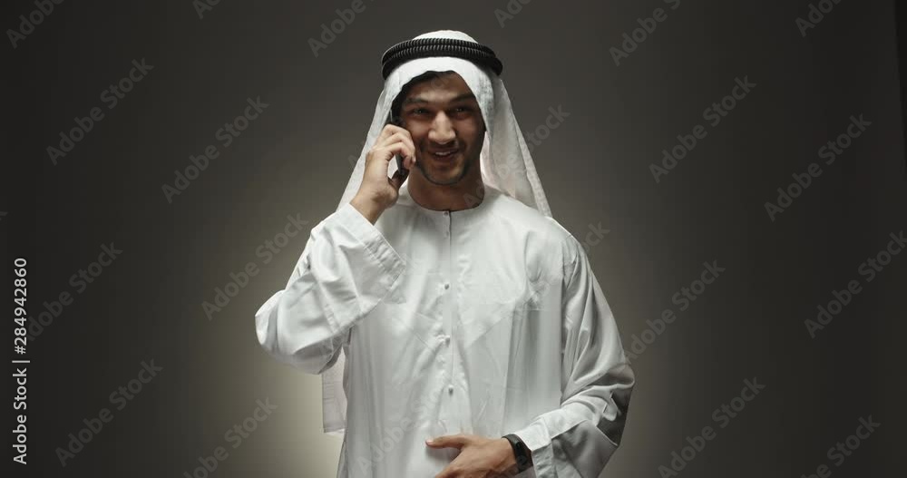 Arabian businessman in traditional white dress talking on phone and positively smiling isolated on black background close up shot 4k Arabian businessman in traditional white dress talking on phone and positively smiling isolated on black background close up shot 4k