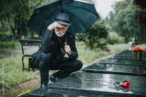 Elegant sad elderly man standing on the rain with umbrella and grieves at the grave of a loved person