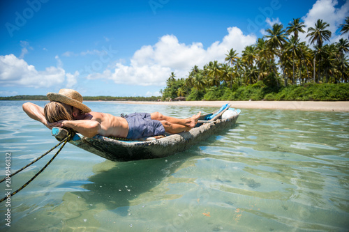 Tourist wearing a straw sun hat lying on a rustic dugout canoe off the shore of a sunny palm-lined tropical island beach in Bahia, Brazil photo