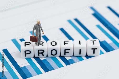 Miniature businessman in suit walking on cube block with alphabets building the word Profit on blue yearly bar chart report, profit, revenue for company or positive return for investment concept