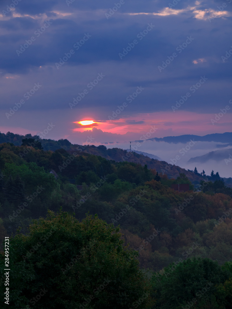 The rising sun peeks from behind heavy rain clouds with a dense fog in the valley.