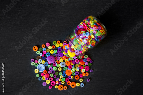 Photos Top view of jar of buttons spilled out onto a black background; colorful buttons