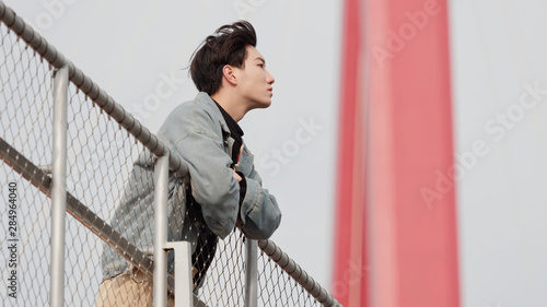 Portrait of a handsome Chinese young man standing and looking away with arms leaning on rail in windy sunny day, enjoy the free wind.