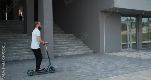 A clear sunny day, an attractive, fashionably dressed man rides scooter along city street, there is no one on sidewalk, bearded man rides calmly and confidently, looking only ahead
