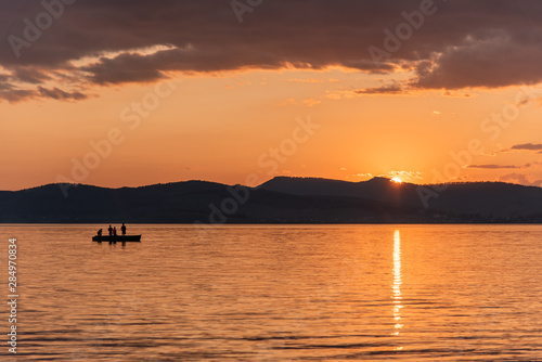 Burning red sunset on the river. Men riding boat. Silhouette of mountains on the background. Beautiful clouds in the sky.