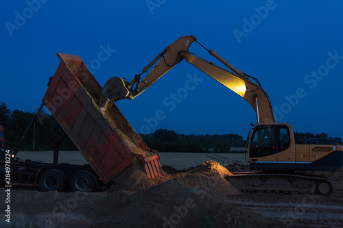 Yellow caterpillar bulldozer unloads a dump truck with sand on the construction of the road. Twilight.