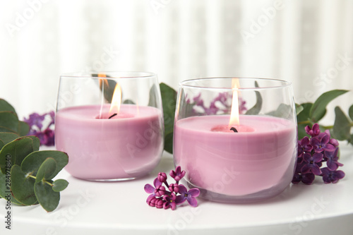 Burning candles in glass holders and flowers with leaves on white table
