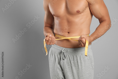 Young man with slim body using measuring tape on grey background, closeup view. Space for text