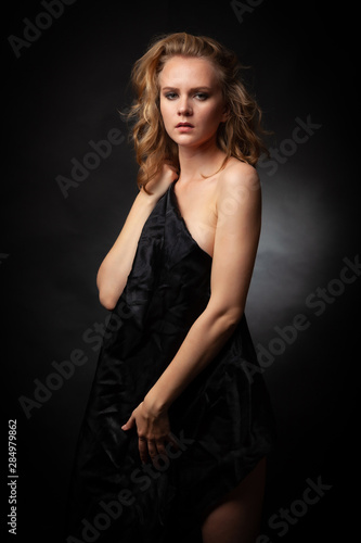 Beautiful naked blonde hiding behind a black cloth stands on a black background.
