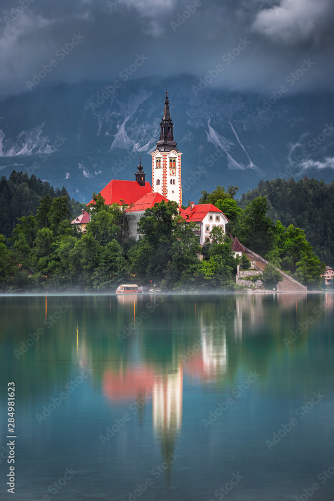 Bled, Slovenia - The Pilgrimage Church of the Assumption of Maria surronded by fog on a small island and Julian Alps covered by clouds taken on a foggy summer morning at Lake Bled (Blejsko Jezero)