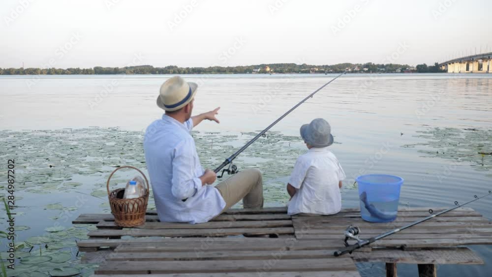 childrens fishing, a cheerful male fisherman teaches his little son how to fish in a river on a fishing rod while sitting on a pier near the water during vacations on a summer evening childrens fishing, a cheerful male fisherman teaches his little son how to fish in a river on a fishing rod while sitting on a pier near the water during vacations on a summer evening