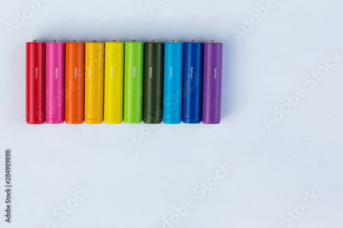 Batteries or accumulators of dimension AA or AAA of all colors of the rainbow lie or stand on a sheet of white paper.