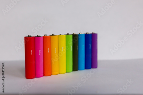 Batteries or accumulators of dimension AA or AAA of all colors of the rainbow lie or stand on a sheet of white paper.