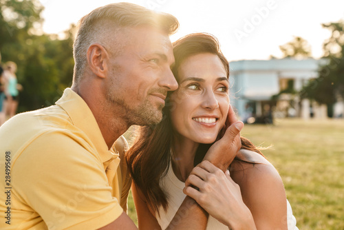 Portrait of lovely middle-aged couple looking aside at copyspace and hugging while sitting on grass in summer park