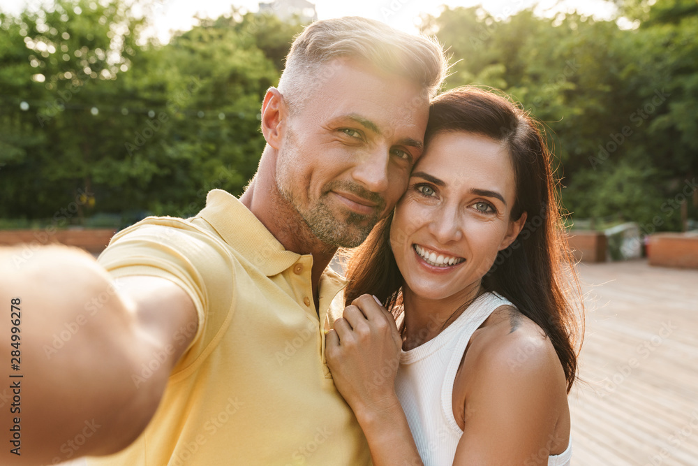 Portrait of happy middle-aged couple taking selfie photo and hugging while walking in summer park Portrait of happy middle-aged couple taking selfie photo and hugging while walking in summer park