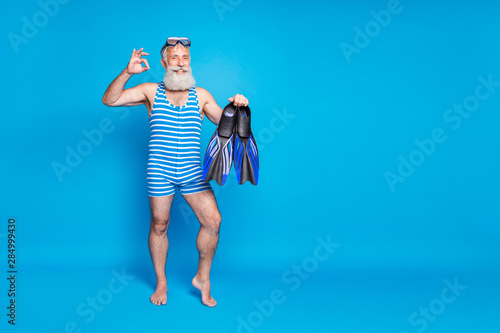 Full length photo of retired pensioner holding fins showing ok sign wearing striped bathing suit isolated over blue background