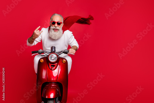Portrait of funky pensioner in eyewear eyeglasses showing rock-and-roll sign driving bike wearing white jumper isolated over red background