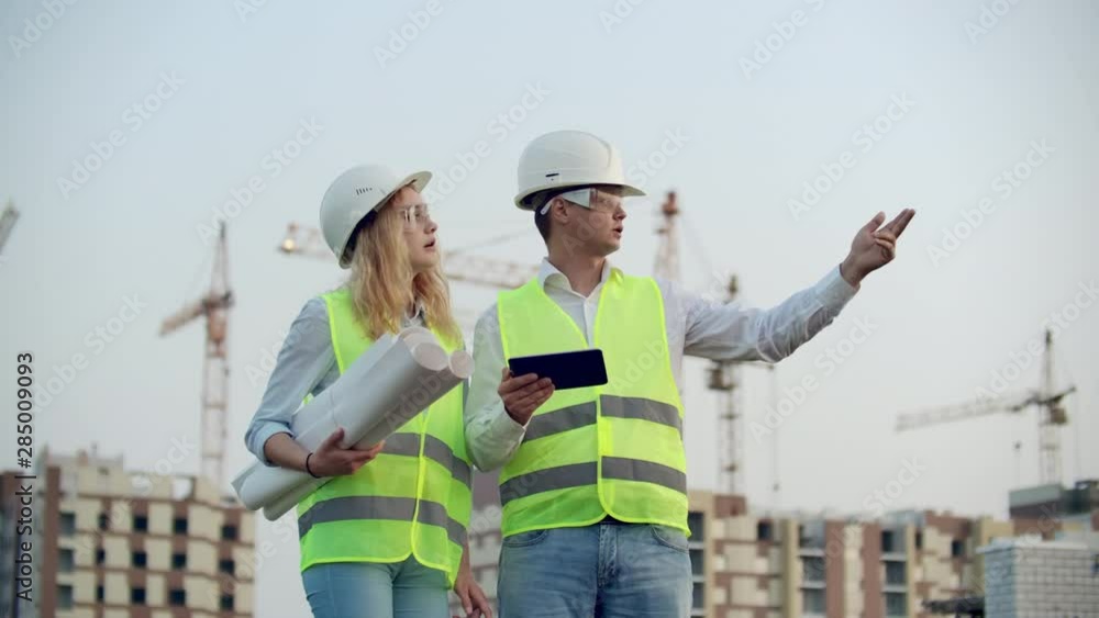 Two colleagues of engineers a man and a woman discussing a drawing and a tablet computer on the background of buildings under construction and cranes, a woman talking on the phone with the boss. Two colleagues of engineers a man and a woman discussing a drawing and a tablet computer on the background of buildings under construction and cranes, a woman talking on the phone with the boss.
