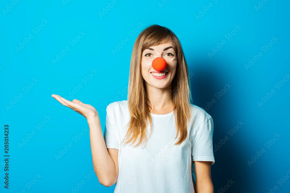 Beautiful young woman in a white T-shirt and a red clown nose smiles and makes hand gestures on a blue background. Concept party, costume, red nose day Beautiful young woman in a white T-shirt and a red clown nose smiles and makes hand gestures on a blue background. Concept party, costume, red nose day