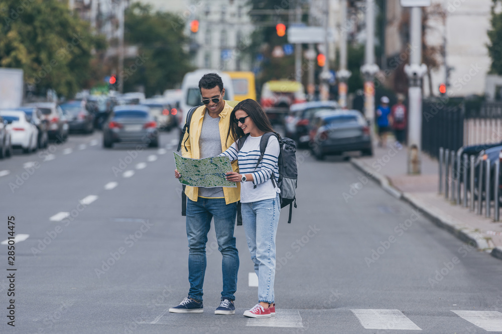 Woman and multiracial friend with backpacks holding city map on crosswalk