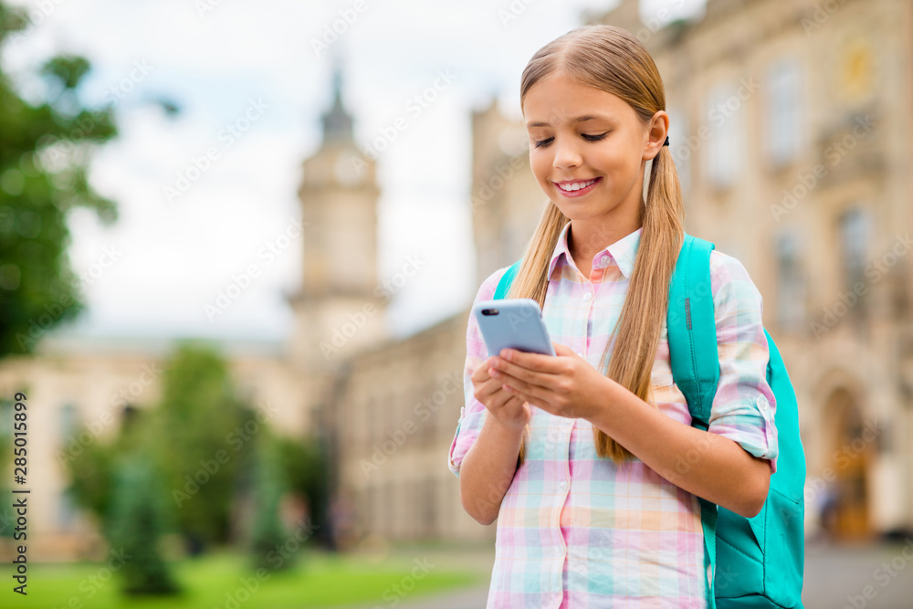 Portrait of concentrated small girl using device holding rucksack backpack wearing checkered plaid t-shirt standing outside Portrait of concentrated small girl using device holding rucksack backpack wearing checkered plaid t-shirt standing outside